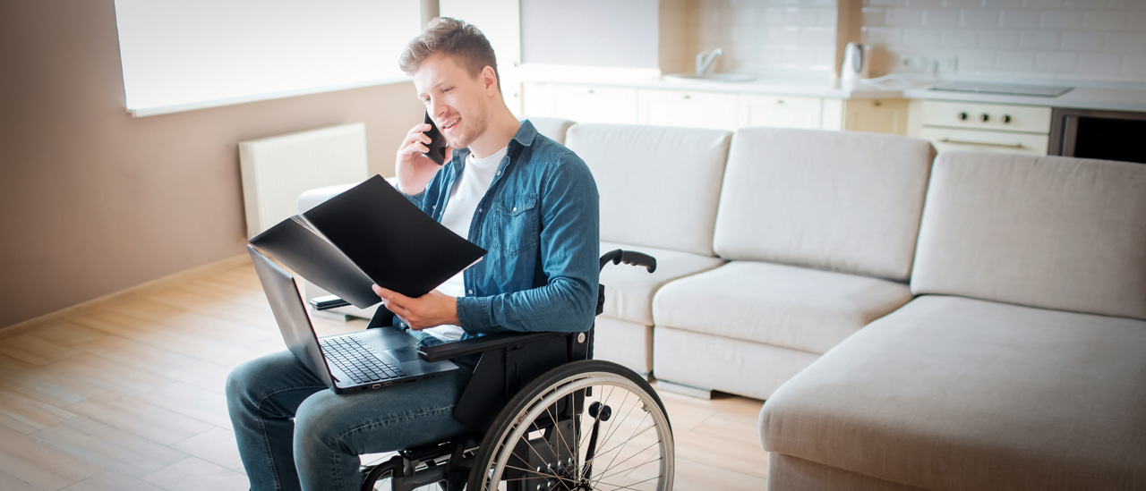 Man, inside his home, sitting in wheelchair looking at an open notebook while talking on the phone.