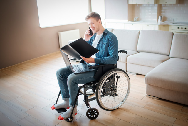 Man, inside his home, sitting in wheelchair looking at an open notebook while talking on the phone.