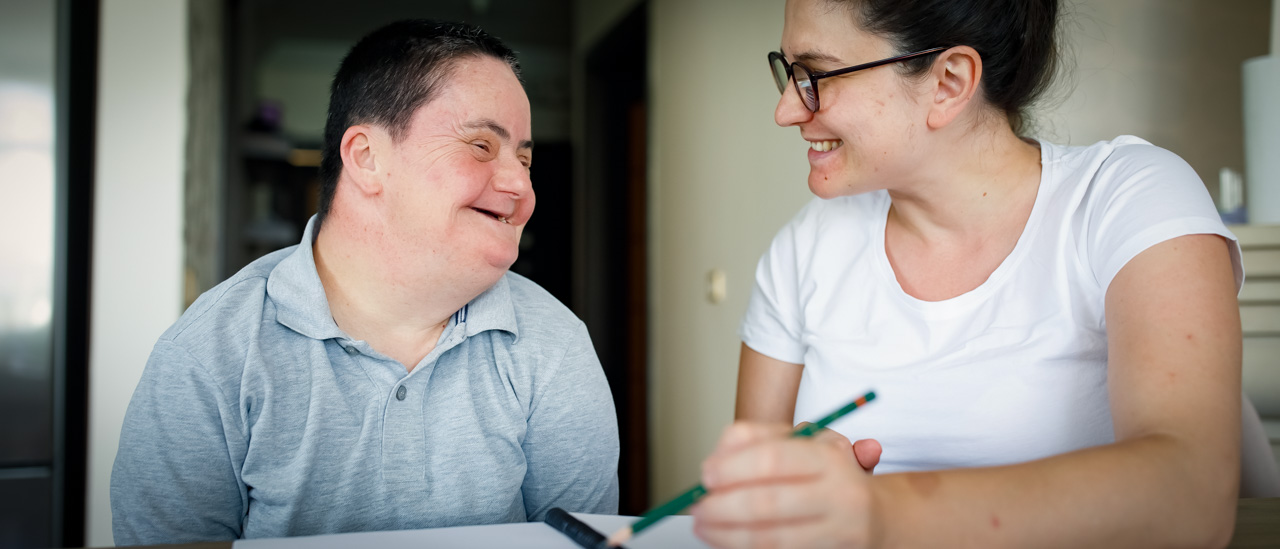 Young man with down syndrome and a woman sitting at a table, talking and taking notes together.