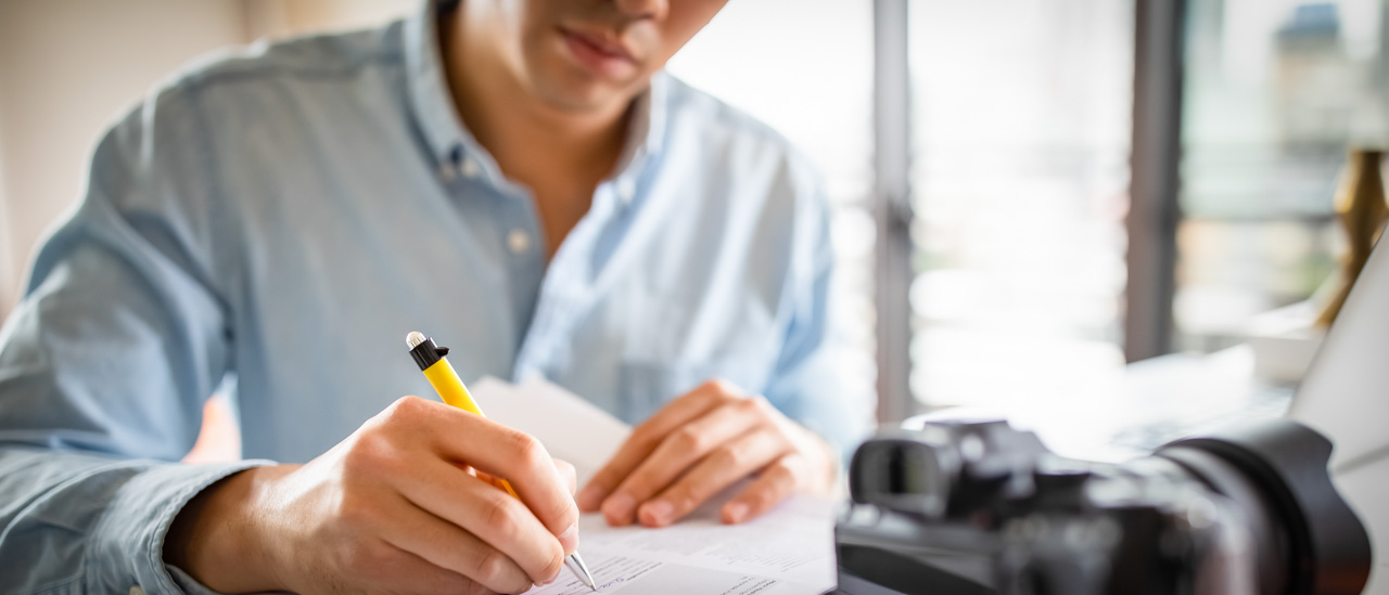 Man at desk notes while his DSLR camera is in front of him.