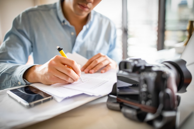 Man at desk notes while his DSLR camera is in front of him.