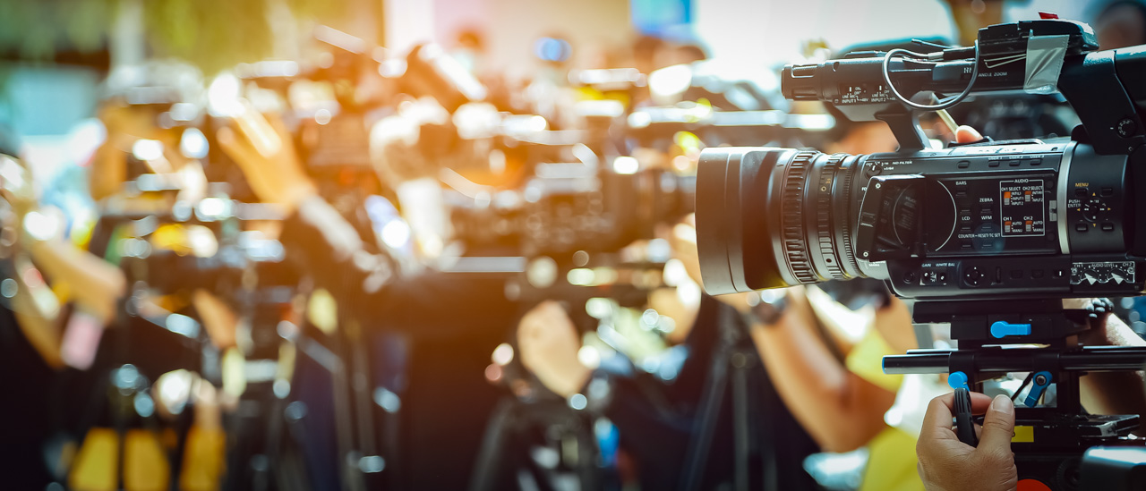 Video camera on front of a blurred group of press and media photographers in the background.