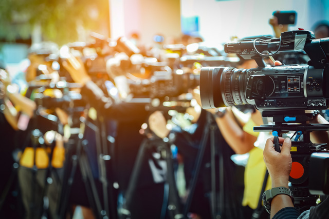 Video camera on front of a blurred group of press and media photographers in the background.