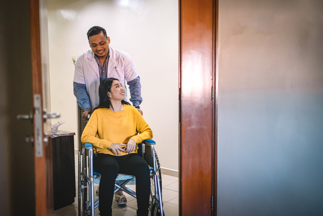 Male nurse talking to a woman while pushing her wheelchair into a hospital room.