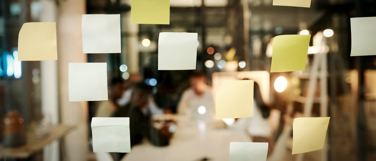 Shot of sticky notes on a glass window with people having a late night brainstorming session in a modern office