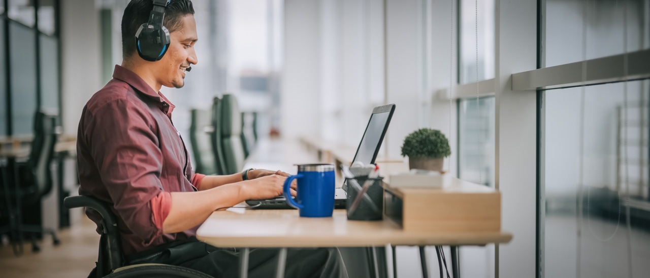 Man wearing headset sitting in wheelchair on front of desk with laptop, having a conversation during an online meeting.