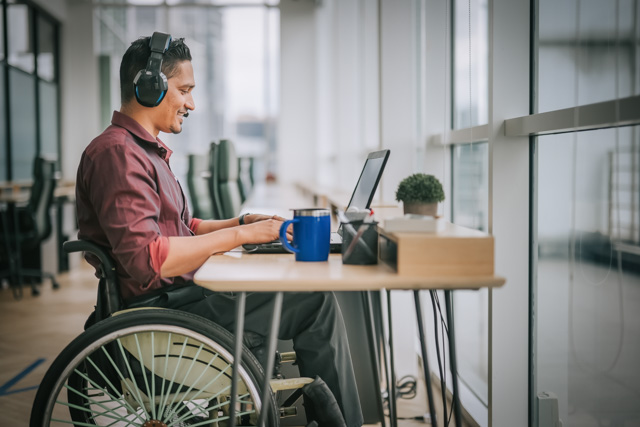 Man wearing headset sitting in wheelchair on front of desk with laptop, having a conversation during an online meeting.