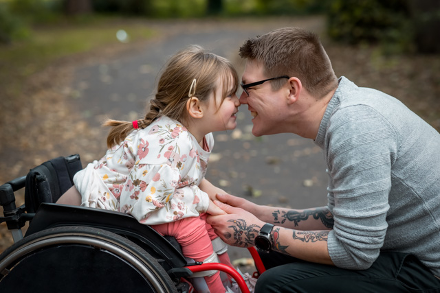 A close up side view of a father and his young daughter who is a wheelchair user having a cute affectionate moment while out in a park.