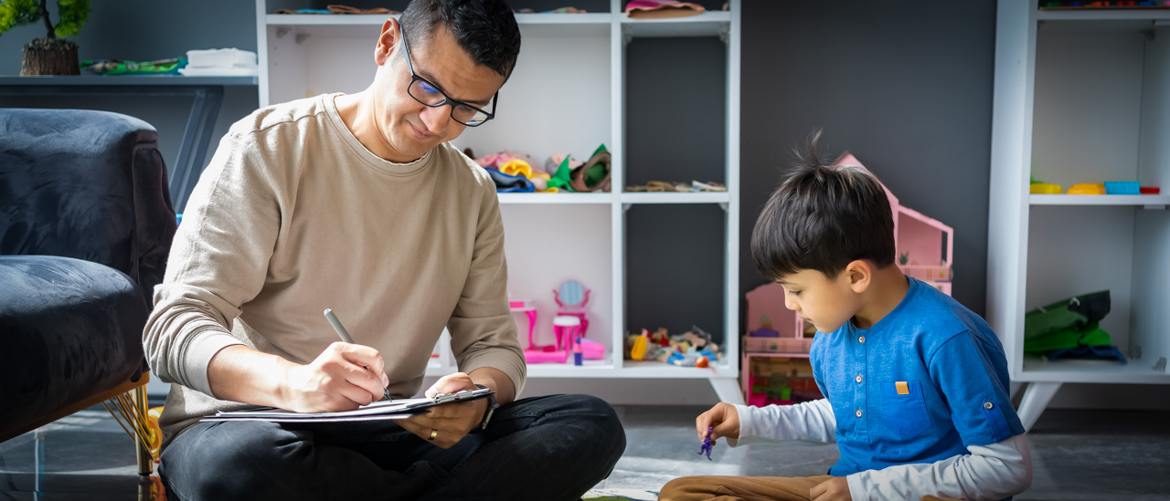 A child psychologist taking notes while observing a boy playing with toys.