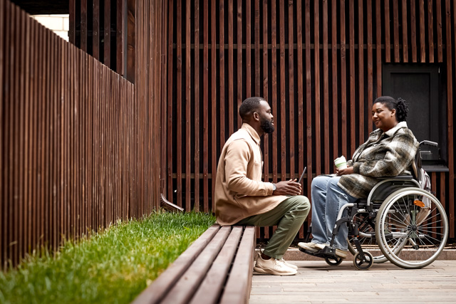 Side view shot of young woman in wheelchair talking to colleague outdoors in city park