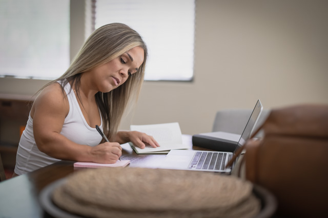 A woman with dwarfism making written notes while using a laptop.