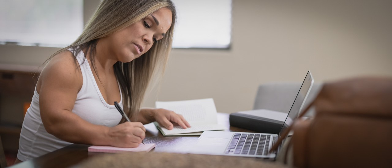 A woman with dwarfism making written notes while using a laptop.