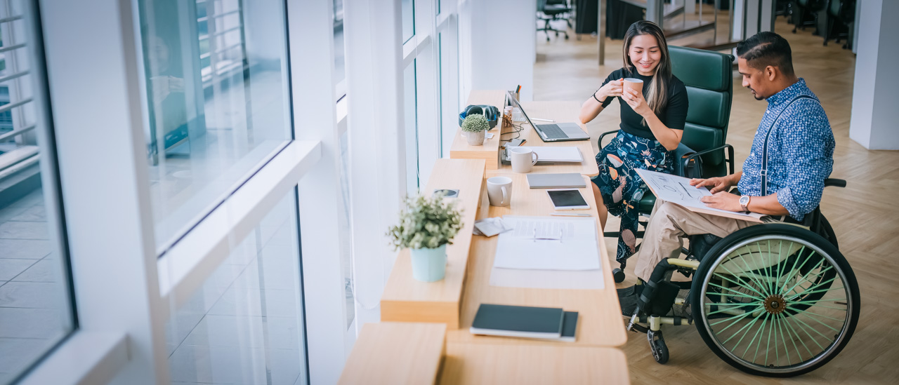 Man in wheelchair and woman sitting next to him having a discussion in an open concept office.