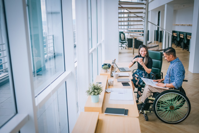 Man in wheelchair and woman sitting next to him having a discussion in an open concept office.