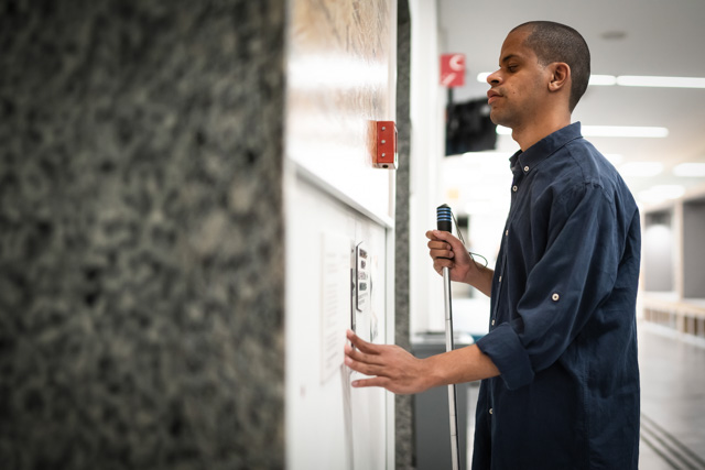 Visually impaired man reading braille on a wall while waiting for an elevator.