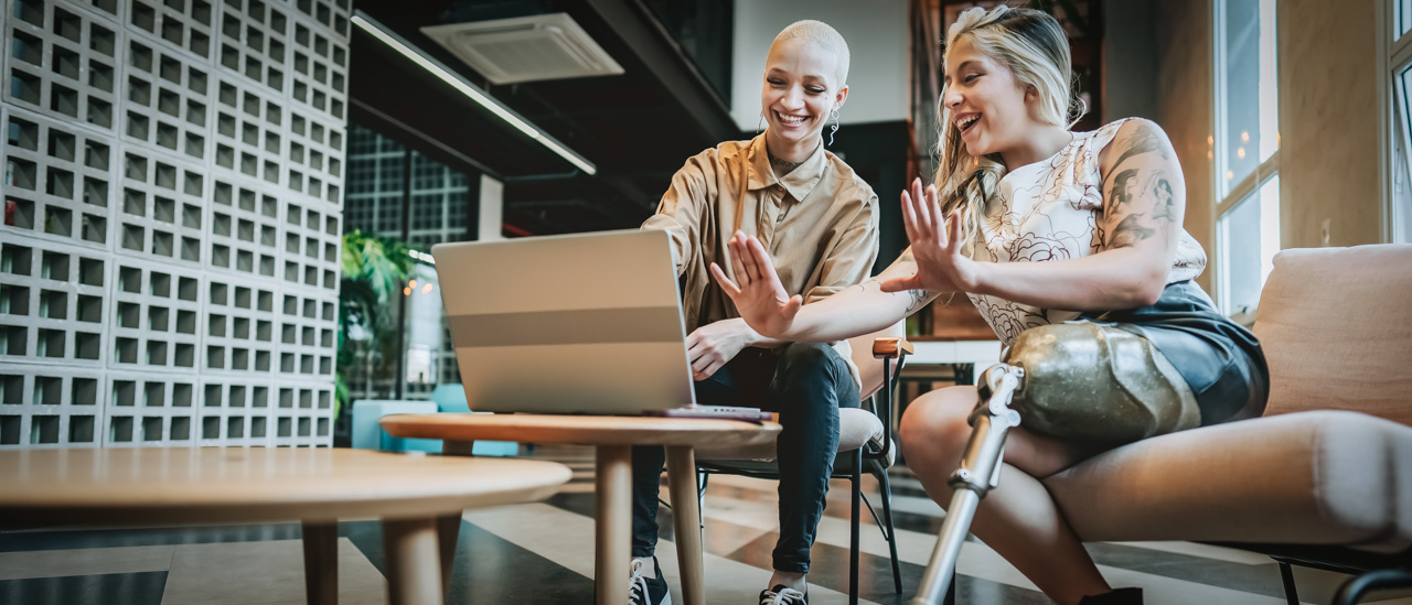 Two smiling women, one with  with a prosthetic leg, sitting on a couch and working on laptop together.