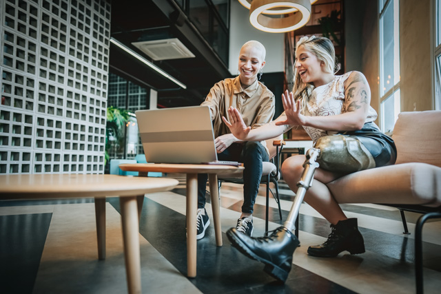 Two smiling women, one with with a prosthetic leg, sitting on a couch and working on laptop together.