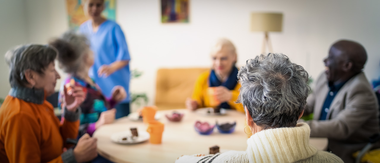 Rear view of an elderly lady sitting in a wheelchair during coffee time with the other residents in a bright modern retirement home.