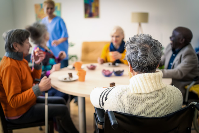 Rear view of an elderly lady sitting in a wheelchair during coffee time with the other residents in a bright modern retirement home.