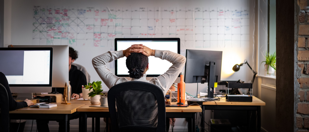 Rear view wide shot of a woman sitting working at a computer in an office with her hands behind her head while looking at her computer screen.