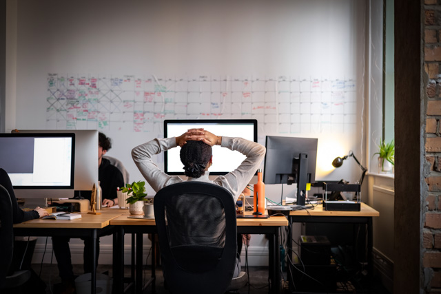 Rear view wide shot of a woman sitting working at a computer in an office with her hands behind her head while looking at her computer screen.