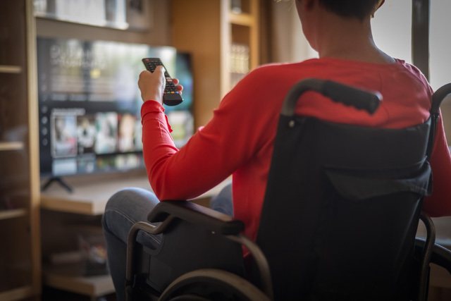 Person in wheelchair holding a remote pointed at a TV in their living room.