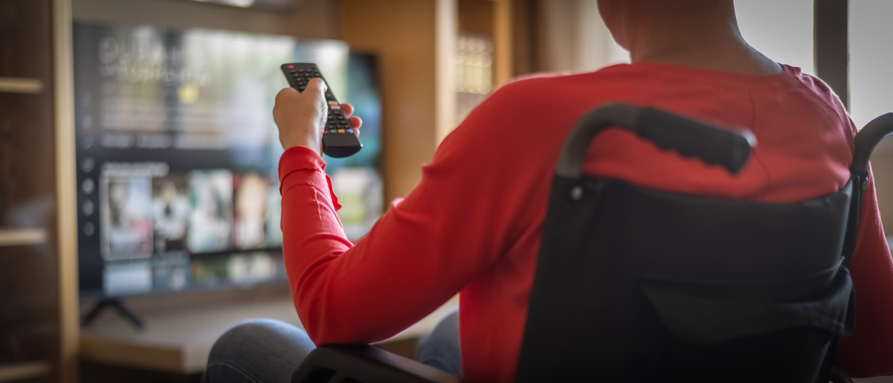 Person in wheelchair holding a remote pointed at a TV in their living room.