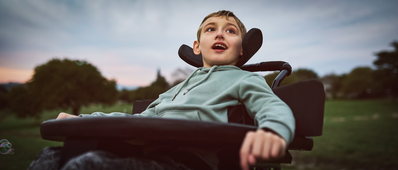 Young boy sitting in wheelchair, out on open field for morning exercise, looking curiously into distance with mouth open.