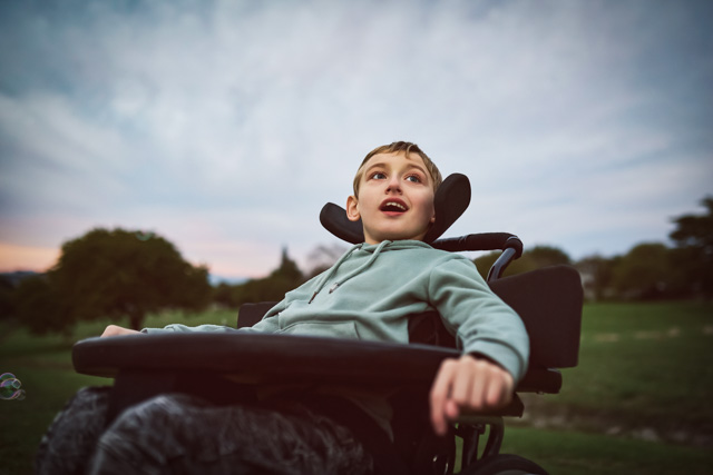 Young boy sitting in wheelchair, out on open field for morning exercise, looking curiously into distance with mouth open.