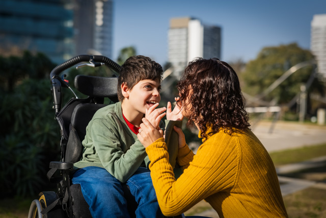 Mother interacting with her son in a wheelchair, playing together in a sunny city park.