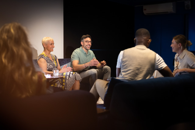 Over the shoulder shot of a group of people inside a dimly lit theatre having a captivating panel discussion. 