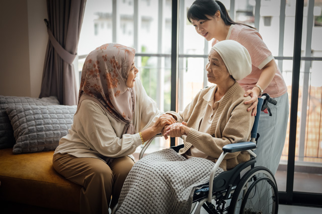 A woman is talking to and holding the hands of her friend seated in a wheelchair, while her friend's caregiver holds on to the wheelchair.
