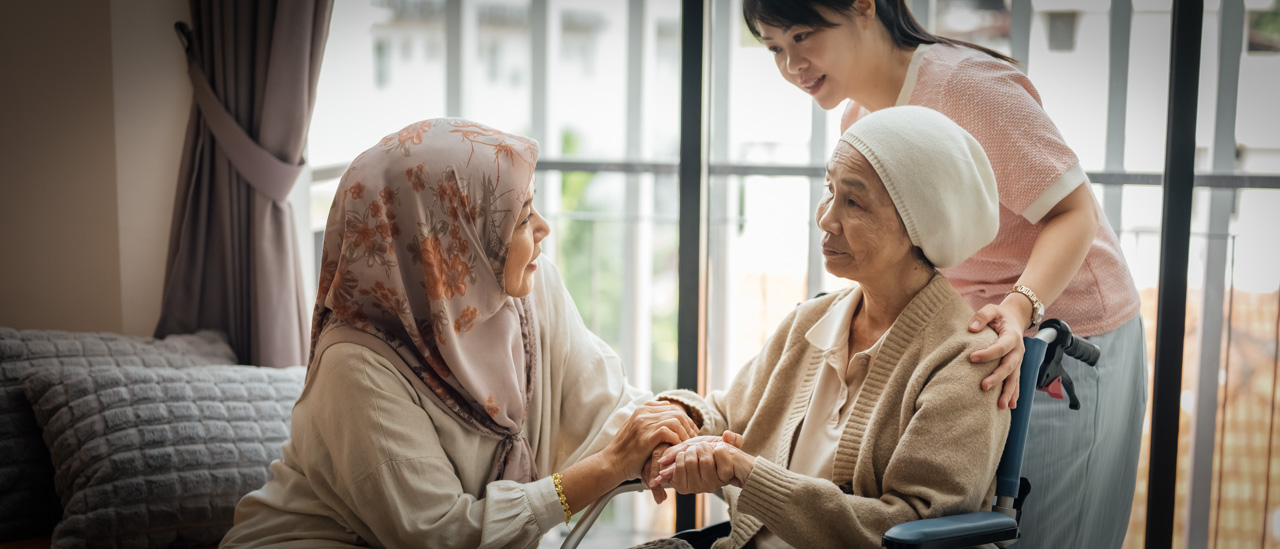 A woman is talking to and holding the hands of her friend seated in a wheelchair, while her friend's caregiver holds on to the wheelchair.