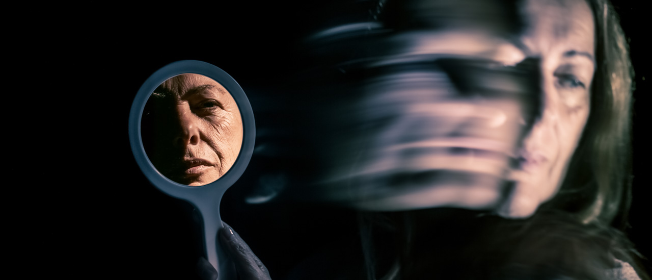 Low light photo of a woman's face blurred and looking away while her is face is clearly reflected in a hand held mirror. 