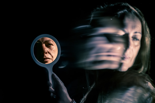 Low light photo of a woman's face blurred and looking away while her is face is clearly reflected in a hand held mirror.